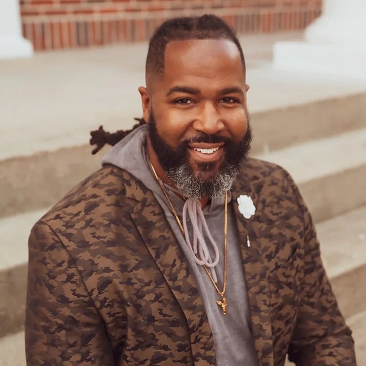 Jordan Brown, smiling Lead Pastor at OV Church, wearing a patterned blazer and gray hoodie, seated on steps, representing community engagement and leadership in church ministries.