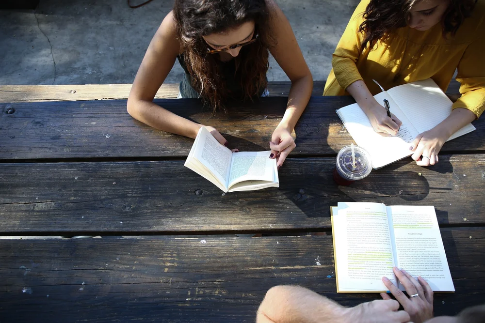 Group studying at a wooden table, one person reading a book, others taking notes in notebooks, emphasizing community learning and engagement.